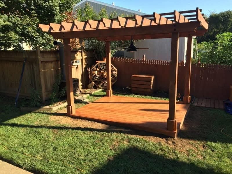 Wooden pergola with benches in a backyard garden on green lawn, surrounded by brown fencing