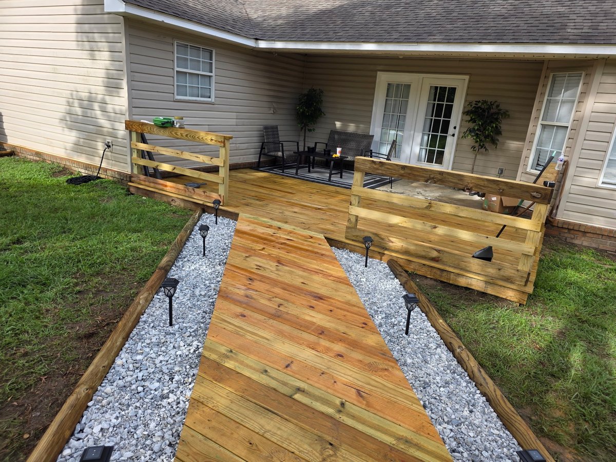 Wooden deck and walkway with white gravel borders leading to a house porch with railings and outdoor furniture