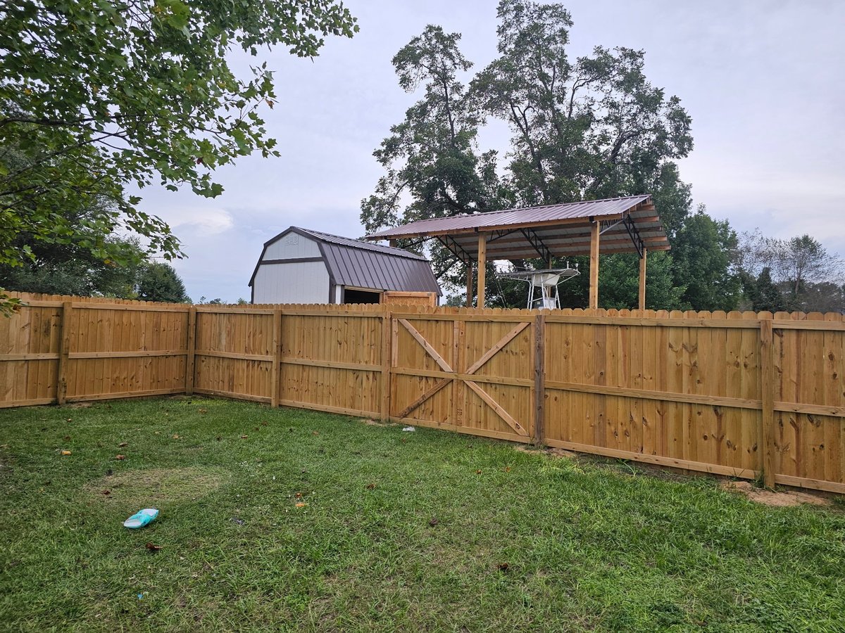 Wooden fenced farmyard with metal barn and open pergola structure, surrounded by large trees and green grass